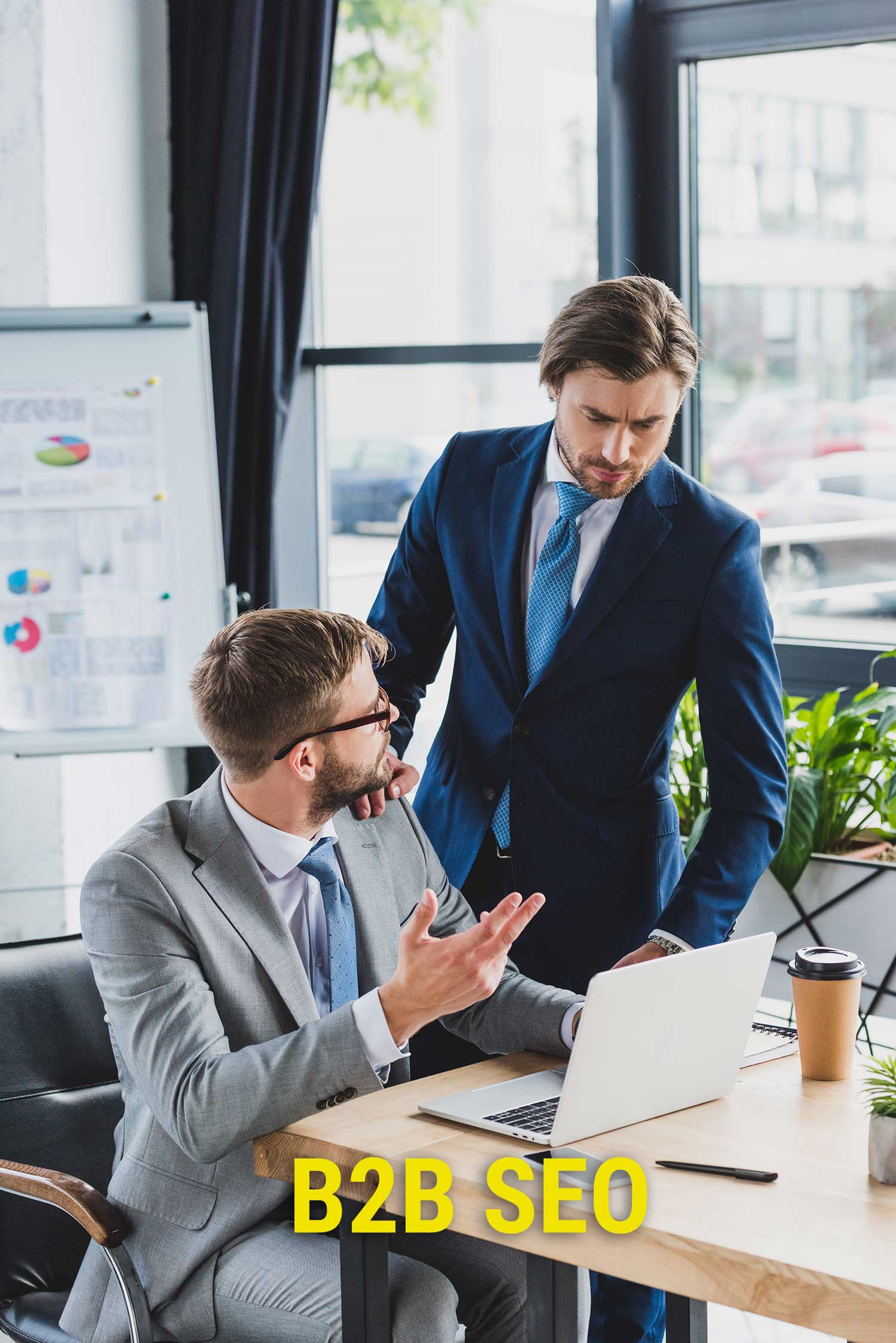 Two businessmen in suits discussing B2B SEO strategy at a laptop in a modern office environment.