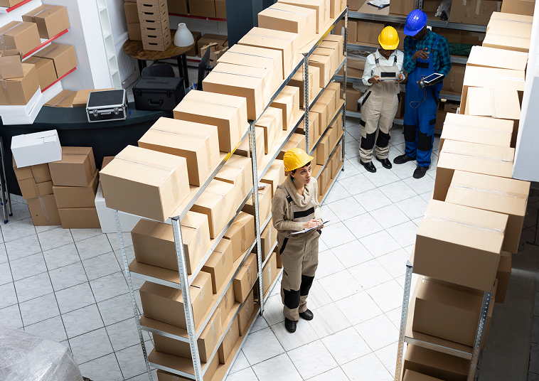 Image shows a wide-angle view of a warehouse with two workers in safety gear and a manager holding a clipboard.