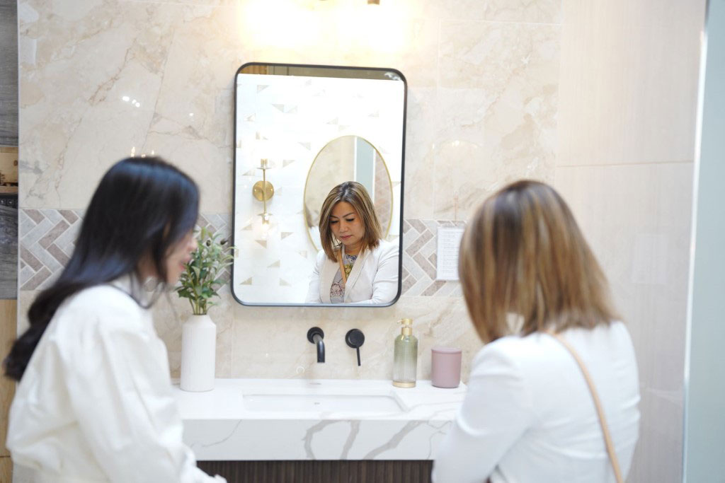 Two women in white jackets examining a bathroom display featuring a white marble vanity, a black faucet, and a rectangular mirror.