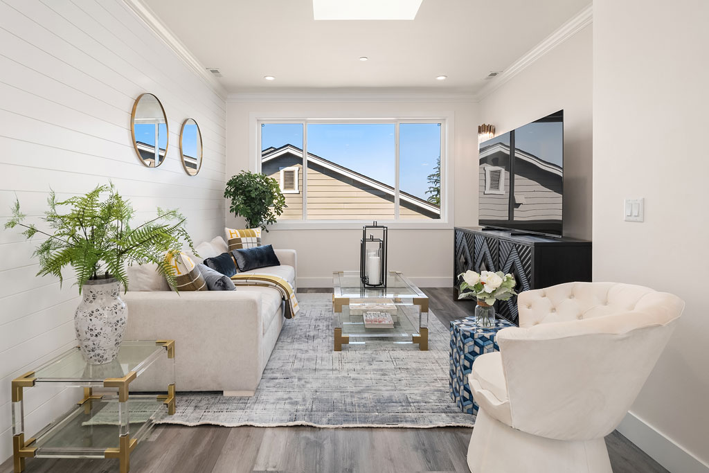 Bright living room with a white shiplap wall, a comfortable white sofa, a glass and gold coffee table, and a large window with a clear sky view.