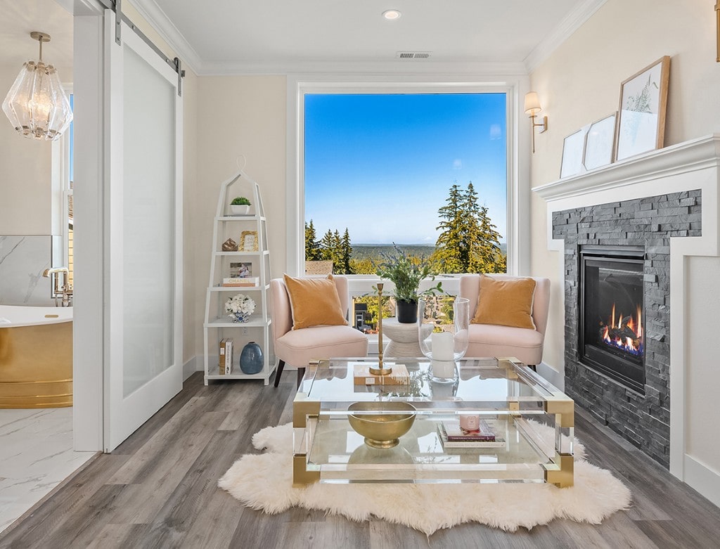 Elegant sitting area with two pink chairs, a glass coffee table, and a stone fireplace, featuring a large window with a scenic view of a forest.