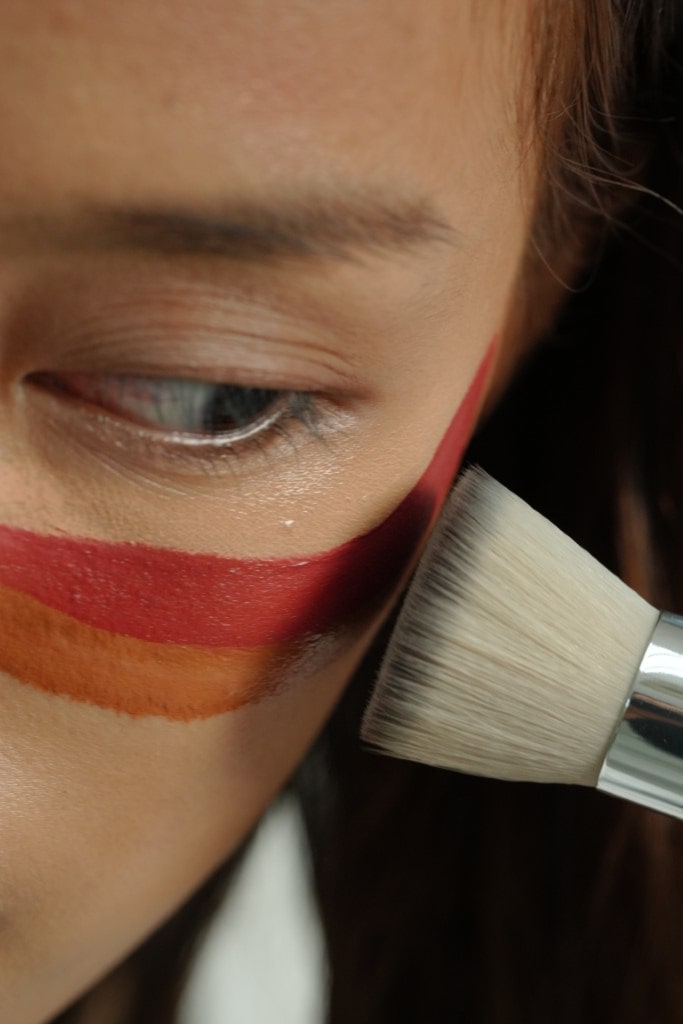 An extreme close-up of a makeup brush blending stripes of red and orange blush on a person's cheek, just below their eye.