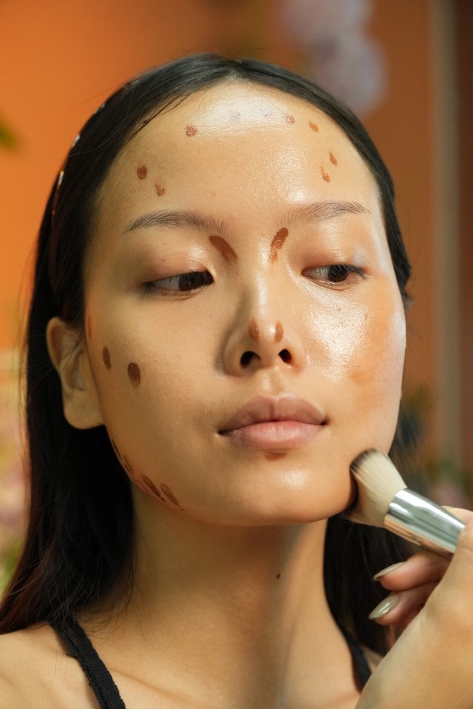 A model with contour makeup applied in dots and lines on her face, with a makeup brush blending the product along her jawline.