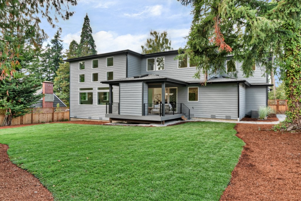 The backyard of a modern two-story home with grey siding, a well-manicured green lawn, a small dark-colored deck, and surrounded by mature trees.