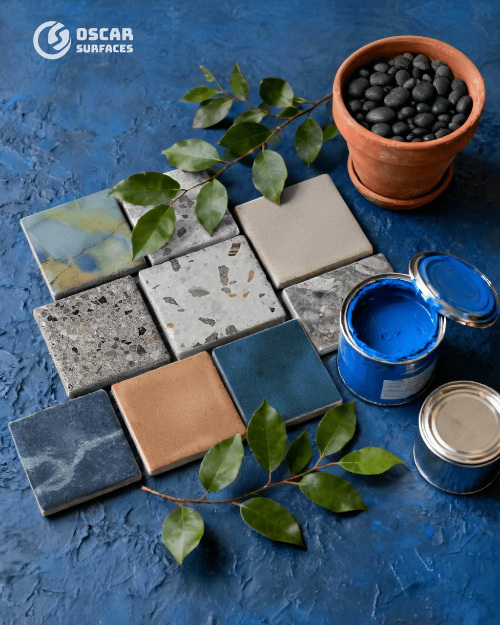 A collection of colorful and textured square sample tiles on a vibrant blue surface, next to an open can of blue paint, a terracotta pot, and green foliage.