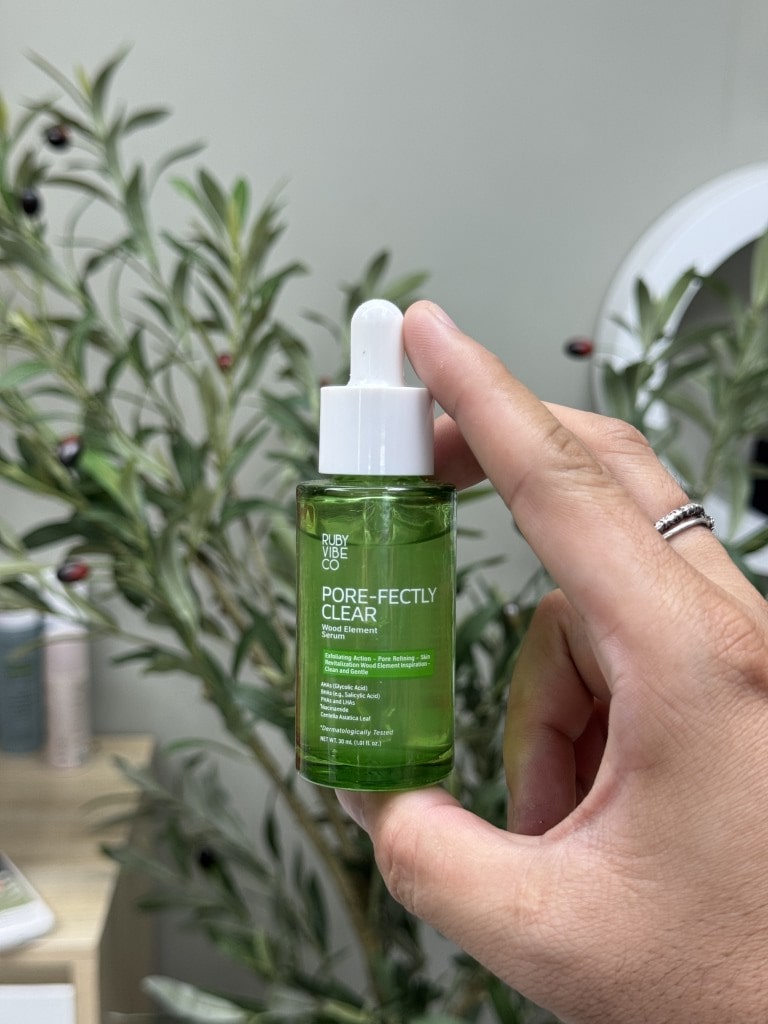 Hand holding small green dropper bottle of "Ruby One Co. Pore-fectly Clear" serum, with olive leaves in background.