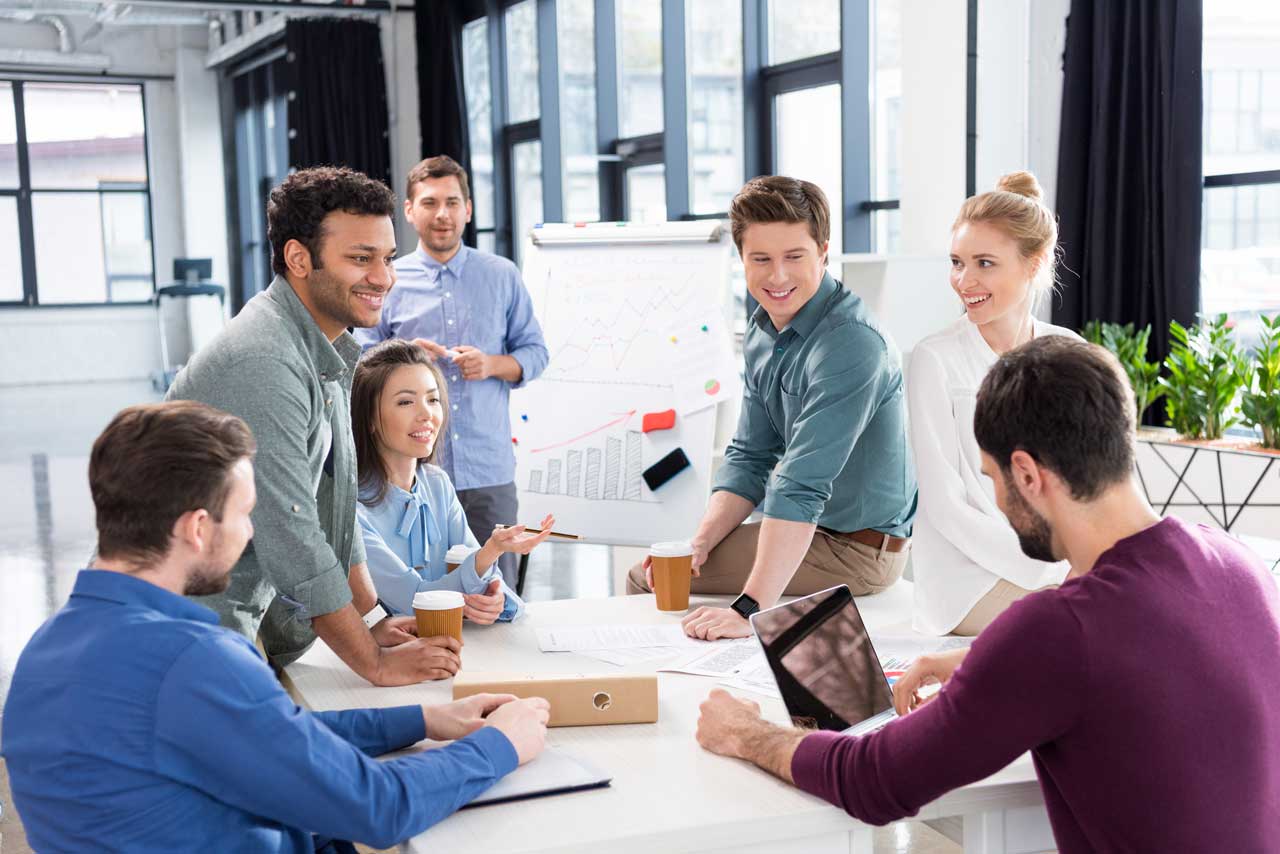 Engaged office team collaborating on B2B sales strategies around a white desk with charts and graphs in the background.