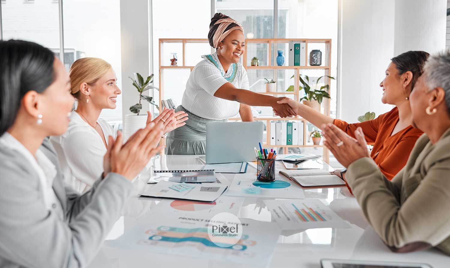 A diverse group of women in a meeting room shaking hands and applauding a business deal.