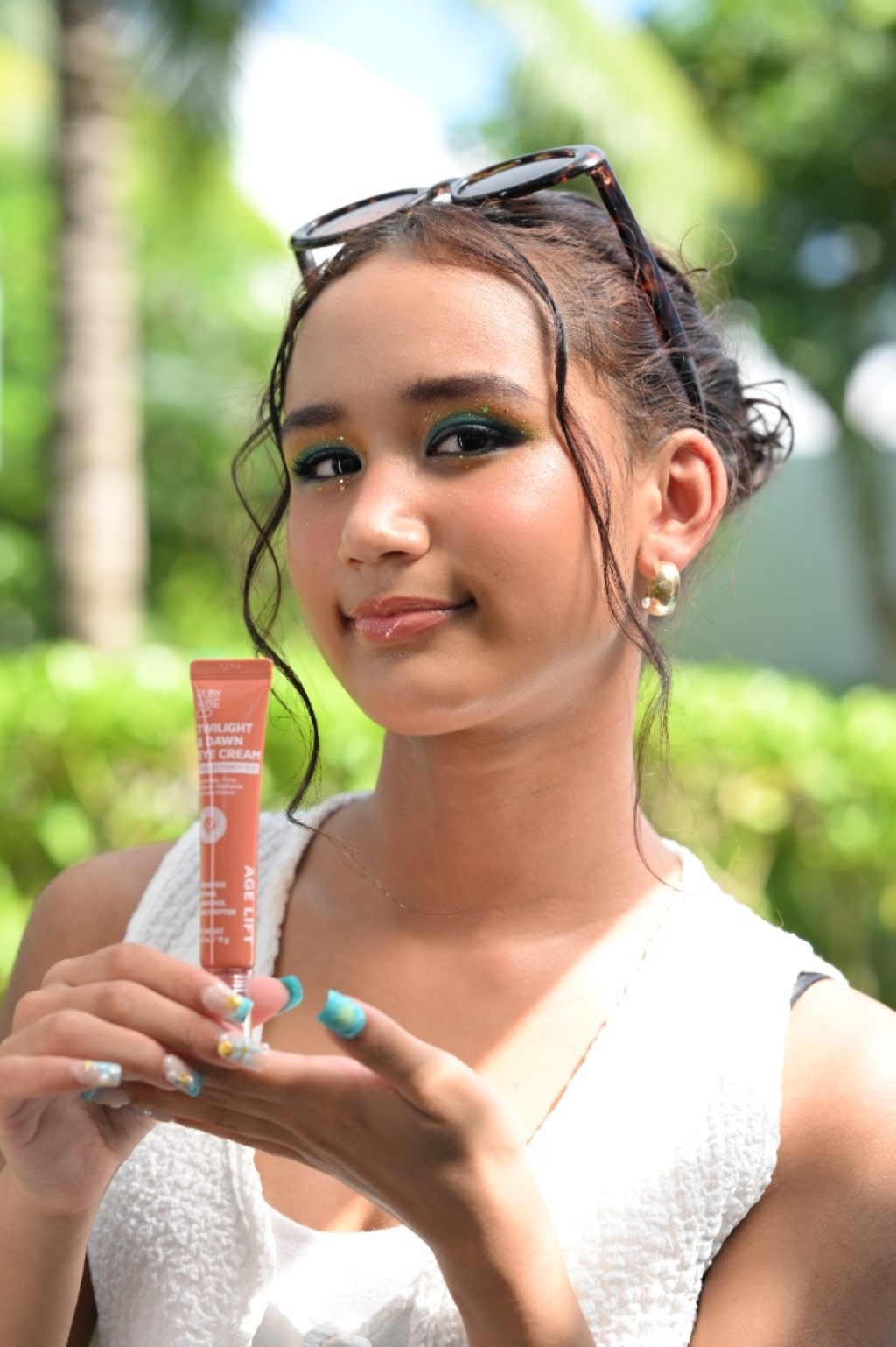 A young woman with sparkling green eye makeup smiles while holding up an orange tube of 'Age Lift' day and night cream, with a lush green background.
